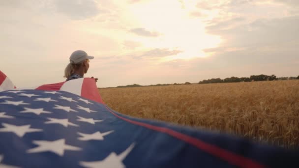 Un granjero patriótico con una bandera de Estados Unidos está cerca de un campo de trigo maduro, sostiene una bandera para uno de los bordes — Vídeo de stock