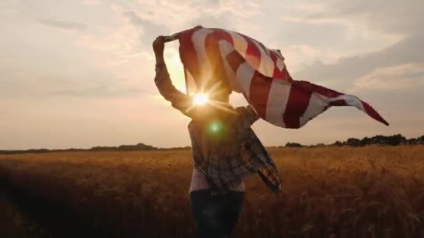Женщина с флагом США бежит на солнце по пшеничному полю — стоковое видео