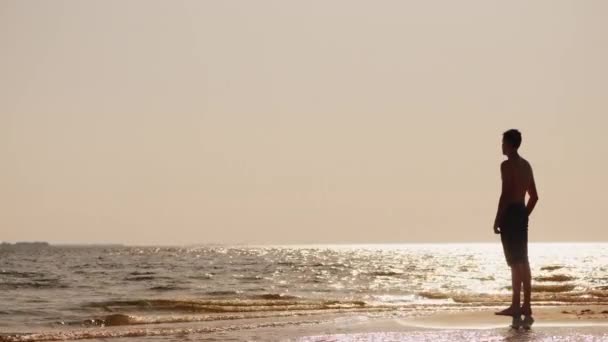 A young man stands on the beach, looking into the distance — Stockvideo