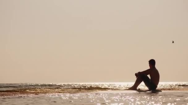 Samotna sylwetka młodego mężczyzny siedzącego na piasku nad morzem — Wideo stockowe