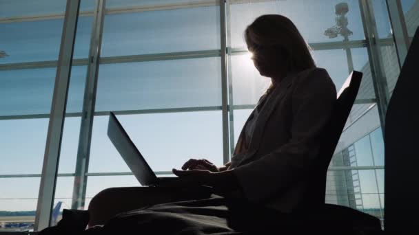 Silhueta de uma mulher de negócios com um laptop, trabalhando em antecipação ao seu voo. Senta-se ao lado da grande janela no terminal do aeroporto — Vídeo de Stock