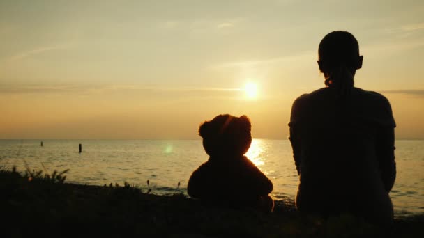 Silhouette of a woman and a teddy bear on the lake, looking forward over the horizon where the sun sets — 비디오