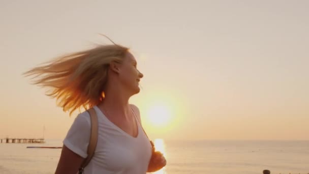 Una donna felice sta camminando felicemente lungo il molo sulla riva del mare con un cappello in mano, ammirando il paesaggio marino la mattina presto. — Video Stock