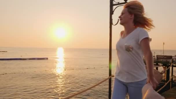 Een gelukkige vrouw loopt vrolijk langs de pier aan de kust met een hoed in haar handen, bewonderend de zeegezicht in de vroege ochtend — Stockvideo