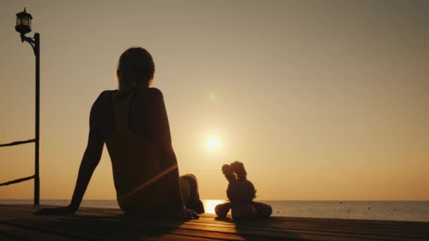 eine Frau mit einem Spielzeugkaninchen sitzt auf einem Steg und bewundert den Sonnenaufgang über dem Meer