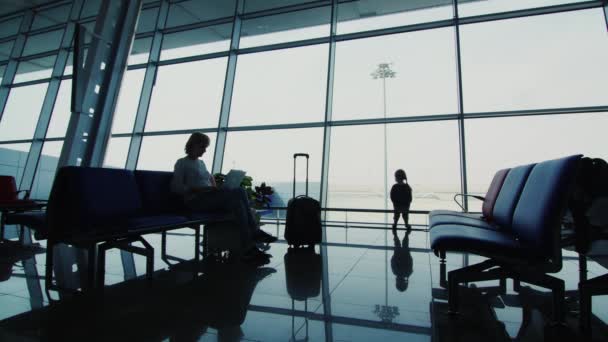 A woman with a child awaiting her flight. Sit on chairs in airport terminal, silhouettes — 비디오