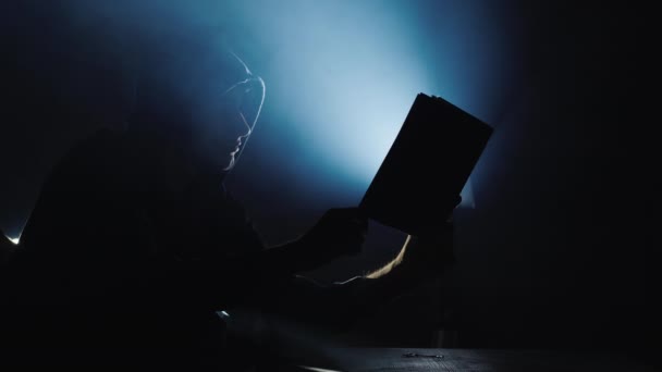 Het silhouet van een man in een kap met een boek in zijn handen, uit het boek komt het licht — Stockvideo