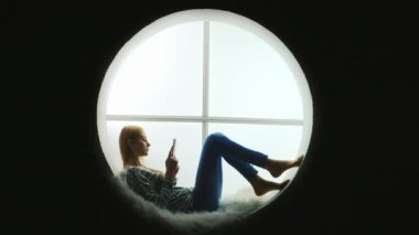 Öğrenci yuvarlak bir pencerede yatıyor, bir e-kitap okuyor.