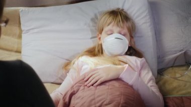 Back view of An elderly woman sits at the bedside of a cold child in a mask, holds the girls hand. Slider shot