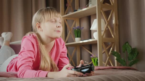 Blond meisje speelt video games op de console, ligt met een gamepad op haar bed — Stockvideo