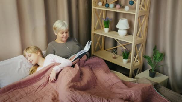 An elderly woman reads a fairy tale to a child who falls asleep nearby — Stockvideo