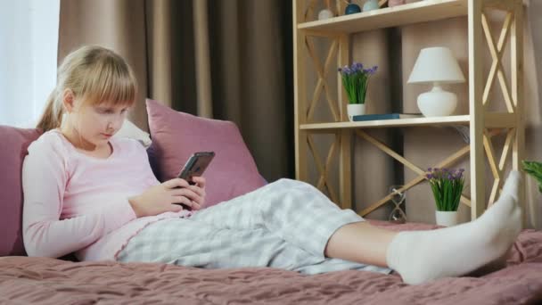 Chica rubia en su cama utiliza un teléfono inteligente — Vídeo de stock