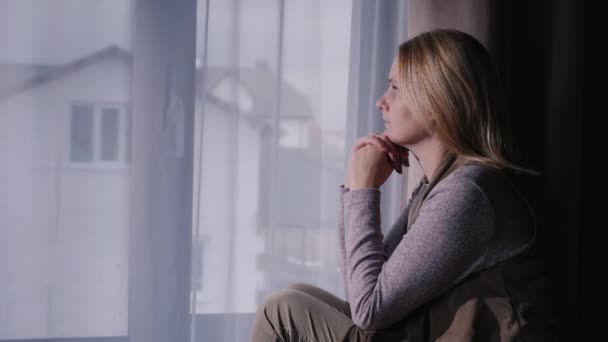 Een trieste vrouw zit op de vensterbank, kijkend uit het raam naar het huis. — Stockvideo