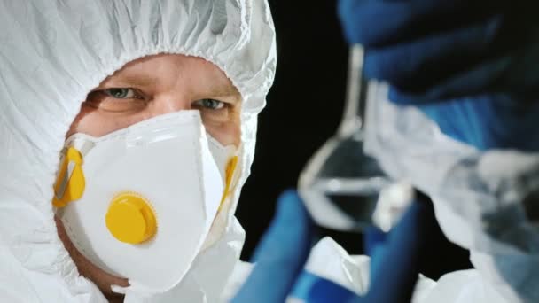 Вчений в захисному костюмі дивиться на пробірку з прозорим препаратом — стокове відео