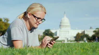 Mutlu kadın Washington 'daki Kongre Binası' nın arka planında çimlerin üzerinde yatıyor. Akıllı telefon kullanıyor..
