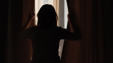 Penceredeki perdeleri açıp ileriye bakan bir kadının silüeti.