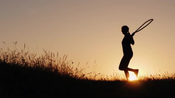 Vista lateral do treinamento ao ar livre - uma silhueta de uma mulher pulando sobre uma corda ao pôr do sol — Vídeo de Stock
