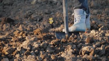 Kadın çiftçi bir sebze bahçesi kazıyor. Çerçevede sadece iş ayakkabılarının bacakları görünüyor.