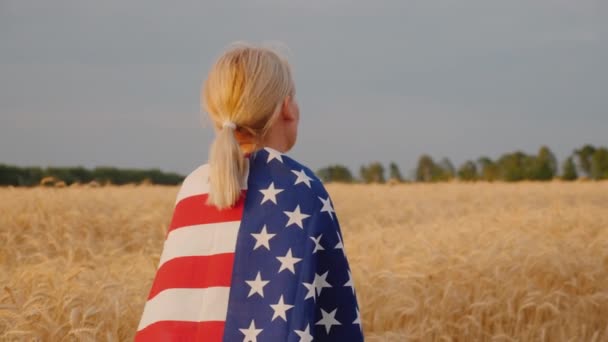 Μια νεαρή γυναίκα με τη σημαία των ΗΠΑ στους ώμους της στέκεται σε ένα χωράφι με ώριμο σιτάρι. — Αρχείο Βίντεο
