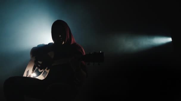 Een jonge muzikant in een capuchon zit op het podium gitaar te spelen — Stockvideo