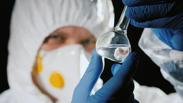 Cientista em terno protetor olha para um tubo de teste com uma droga transparente — Fotografia de Stock