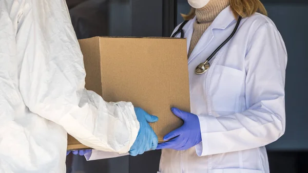 Koruyucu giysili bir kişi hastanenin girişindeki doktora bir kutu ilaç veriliyor.
