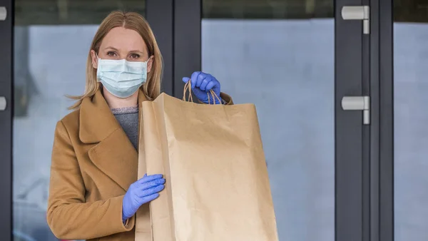 Eldivenli ve maskeli bir kadın markete girerken arka planında torbalar dolusu yiyecek olan.