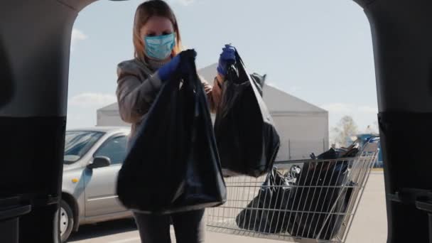 Uma mulher com uma máscara e luvas coloca sacos de compras no porta-malas de um carro — Vídeo de Stock