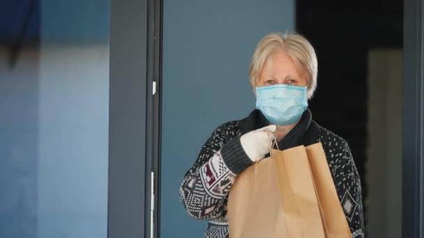 Das Porträt einer älteren Frau mit Maske, die Lebensmittelbeutel in der Hand hält. Hauszustellung in Quarantäne — Stockvideo