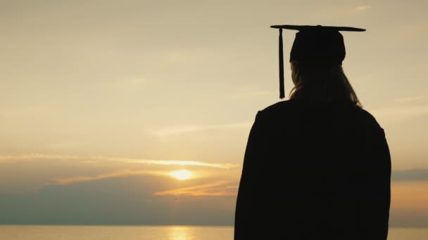 Una mujer con una bata de graduado y una gorra mira el amanecer sobre el mar — Vídeos de Stock