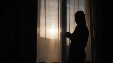 Penceredeki siluet kadın, sıcak bir içecek içer ve doğan güneşe bakar.