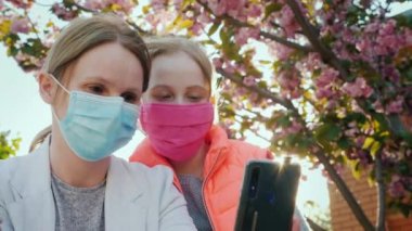 Koruyucu maskeli anne ve kızı, kiraz çiçeklerinin arka planında kendi resimlerini çekiyorlar.