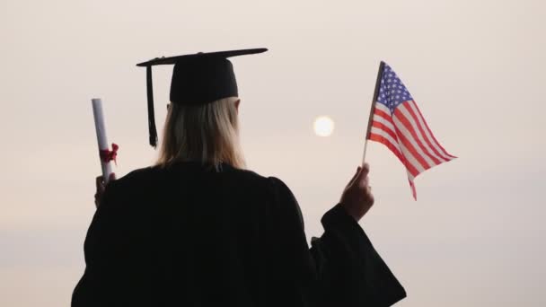Студент в мантии и кепке имеет диплом и флаг США. Учеба в США — стоковое видео