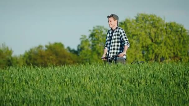 Smiling young man walks with his bike through the picturesque terrain among the fields of wheat. — Stock Video