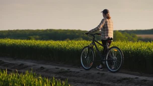 Een vrouw leidt een fiets langs de groene velden van tarwe, geniet van een ontspannen wandeling — Stockvideo