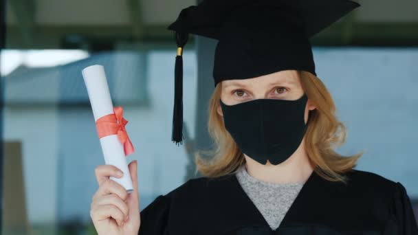 Een vrouwelijke afgestudeerde in een masker houdt een diploma in haar hand — Stockvideo