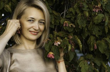 Bir yaz parkında gülücükler saçan orta yaşlı sarışın bir kadın. Bir ağacın arka planında ve yeşil yapraklarda.