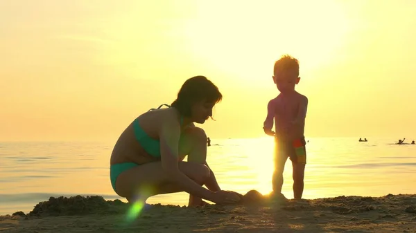 Happy mom and child playing on the beach in the sand. Mom and child build a sand castle against the background of a sea sunset. The concept of a happy family and rest.