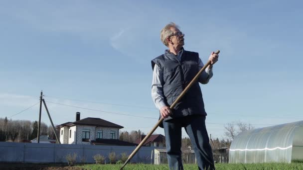 Un uomo anziano con gli occhiali e un giubbotto blu è in piedi con un rastrello in mano . — Video Stock
