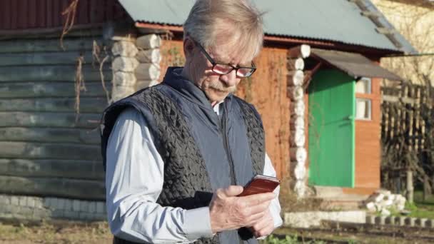 Starszy mężczyzna w okularach i niebieskiej kamizelce wybiera informacje na smartfonie. — Wideo stockowe