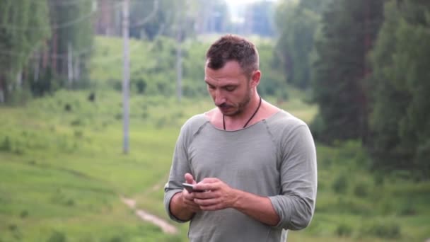 Un joven se para en el fondo del bosque y mira el teléfono . — Vídeos de Stock