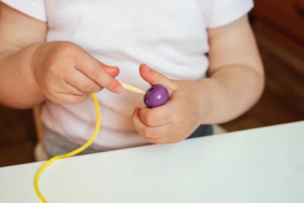 Çocuk ipin ucuna boncuk takıyor. Boncuk dizme aktivitesi. Güzel motor beceri geliştirme. Erken eğitim, Montessori Metodu. Bilişsel beceriler, çocuk gelişimi. Bebeğin ellerini kapayın. Bağlama, iplik etkinlikleri. 