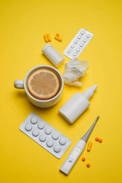 Pills, medical thermometer, nose spray, lemon tea. Different medications on yellow background. Cold and flu home remedies. Disease self-treatment concept. Flat lay. Vertical.
