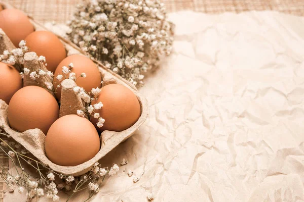 A tray of brown fresh hen\'s eggs decorated with flowers on wrinkled craft paper. Eco-friendly egg production. Bakery ingredients. Organic chicken eggs. Eco Easter concept.  Free space.