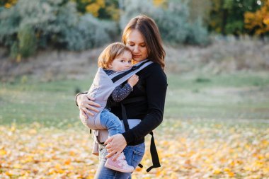 Genç bir anne askısında bebeğini taşıyor. Ailesi olan bir çocuk. Taşıyıcıdaki küçük tatlı kız. Eklenti ebeveynliği kavramı.