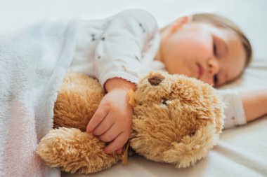 Küçük bebek beyaz çarşaflarda uyuyor yumuşak kahverengi oyuncak ayıya sarılıyor. Çocukların uyku zamanı ya da yatma zamanı. Çocuk uykusuzluğu. Çocuk uyku rutini ve program. Kapat..