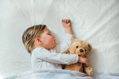 Küçük bebek beyaz çarşaflarda uyuyor yumuşak kahverengi oyuncak ayıya sarılıyor. Çocukların uyku zamanı ya da yatma zamanı. Çocuk uykusuzluğu. Çocuk uyku rutini ve program. Üst görünüm. 