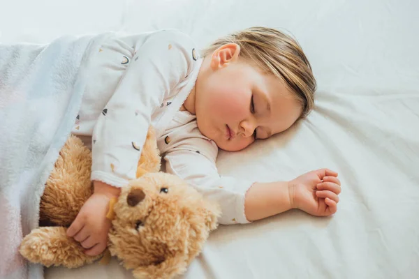 Küçük bebek beyaz çarşaflarda uyuyor yumuşak kahverengi oyuncak ayıya sarılıyor. Çocukların uyku zamanı ya da yatma zamanı. Çocuk uykusuzluğu. Çocuk uyku rutini ve program.