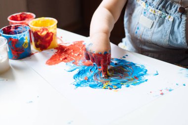 Evdeki masada ellerini mavi ve kırmızı boya kullanarak resim yapan bir çocuk. Çocuklar için parmak boyama ya da sanat terapisi. Bebekler için eğlenceli aktiviteler. Kapat.. 