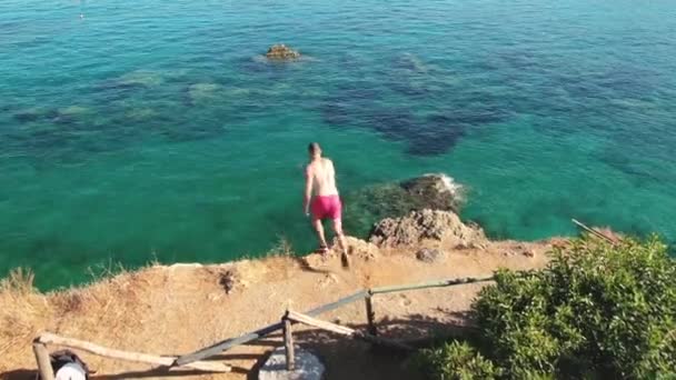 Homem aéreo corre para cima e salta de um penhasco para o mar azul em câmera lenta, salpicos voar em direções diferentes. Divertir-se nas férias de verão . — Vídeo de Stock